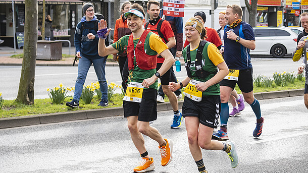Zwei Läufer:innen beim Marathon, wovon eine Person freudig in die Kamera winkt.