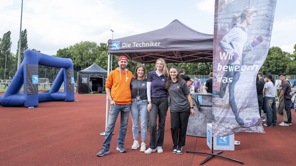Man sieht drei Frauen und einen Mann die nebeneinander auf einem Sportplatz stehen und in die Kamera lächeln. Im Hintergrund ist ein Pavillon mit dem Logo der Techniker Krankenkasse aufgebaut und eine Fahne der Techniker Krankenkasse. Im Hintergrund steht eine Gruppe von Menschen.