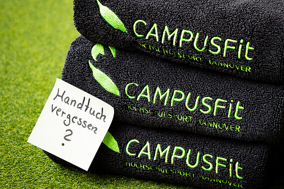 Das Bild zeigt zwei Handtücher, die mit dem Hochschulsport-Logo versehen sind. Auf ihnen klebt ein Post-It mit der Aufschrift: "Handtuch vergessen?".