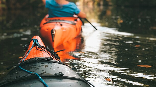 Blickwinkel als Kajakfahrer auf eine Kajakfahrerin vor dem eigenen Kajak auf einem Fluss.