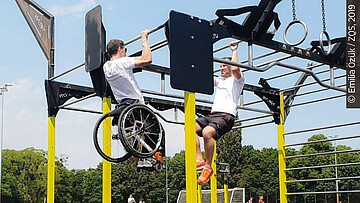 Zwei Personen, eine davon im Rollstuhl, trainieren am Outdoor-Rack.