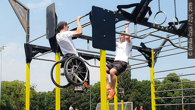 Zwei Personen, eine davon im Rollstuhl, trainieren am Outdoor-Rack.