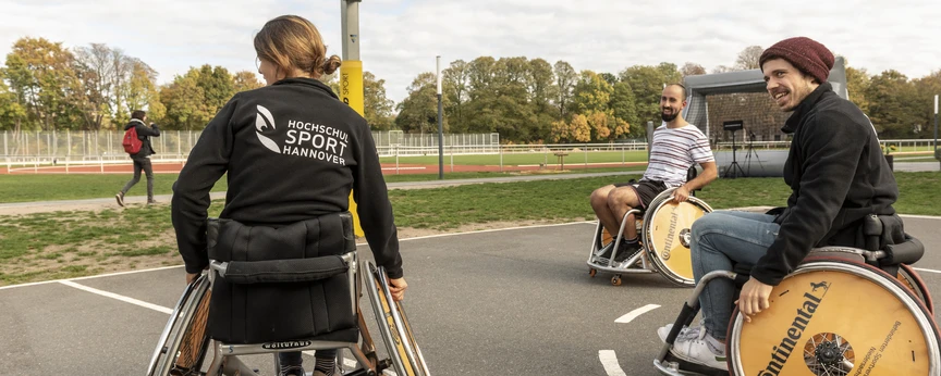 Eine Gruppe Menschen spielt Rollstuhl Basketball. Auf einer Sweatjacke erkennt man das Logo des Zentrums für Hochschulsport.