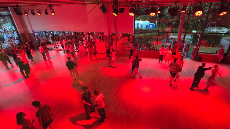Eine Sporthalle wird mit roten Deckenscheinwerfern beleuchtet. Männlich und weiblich gelesene Personen tanzen Paartanz in der Sporthalle.