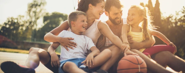 Familie mit zwei Kindern sitzt gemeinsam auf einem Basketball Court.