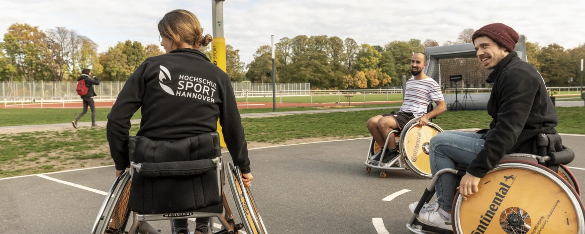 Eine Gruppe Menschen spielt Rollstuhl Basketball. Auf einer Sweatjacke erkennt man das Logo des Zentrums für Hochschulsport.