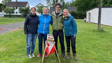 Ein Mann und eine Frau schauen lächelnd in die Kamera. Die Frau hält eine Urkunde in der Hand. Im Hintergrund ist ein Boxring zu sehen. Bild 2: Drei Frauen und ein Mann stehen nebeneinander auf einer Rasenfläche und lächeln in die Kamera. Im Hintergrund sind Bäume und Häuser zu sehen.