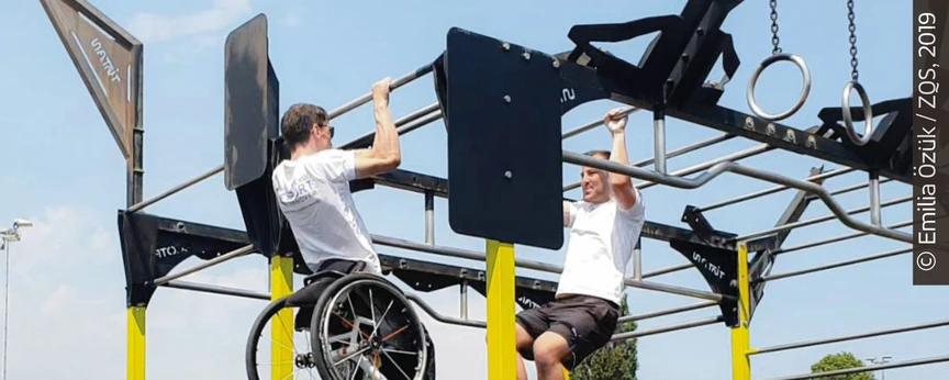 Zwei Personen, eine davon im Rollstuhl, trainieren am Outdoor-Rack.